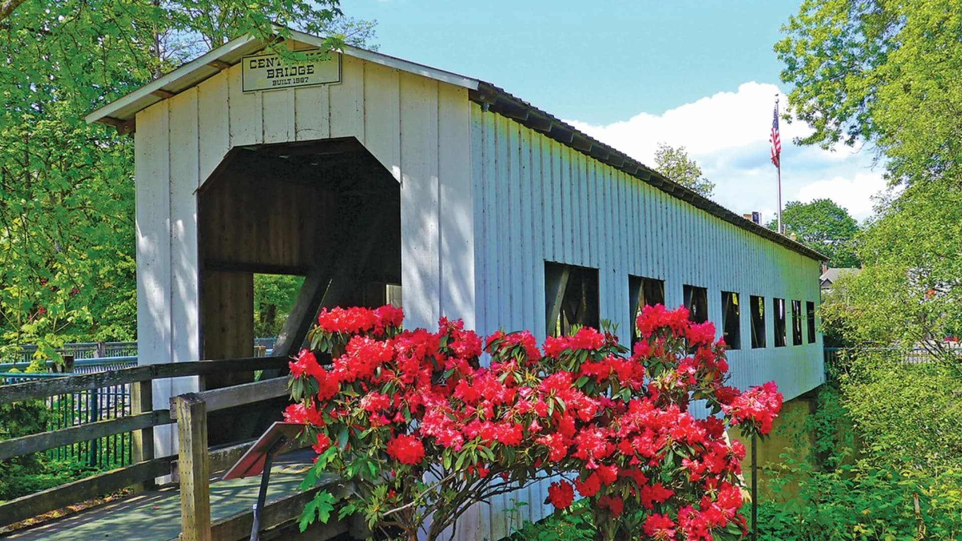 Covered bridge named "Centennial Bridge" surrounded by vibrant red flowers and green foliage.