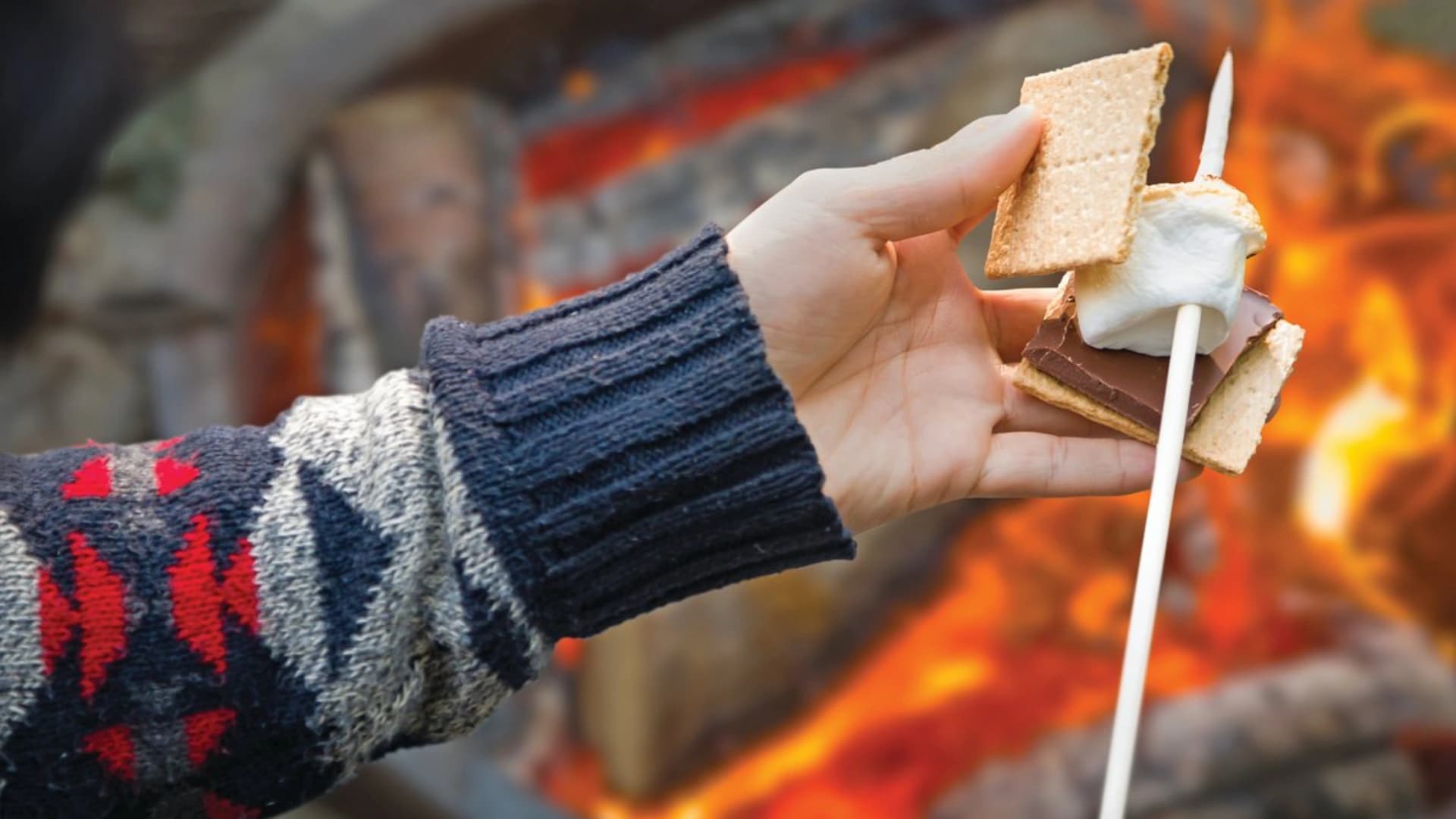 A hand holding a s'more with graham crackers, chocolate, and a marshmallow in front of a campfire.