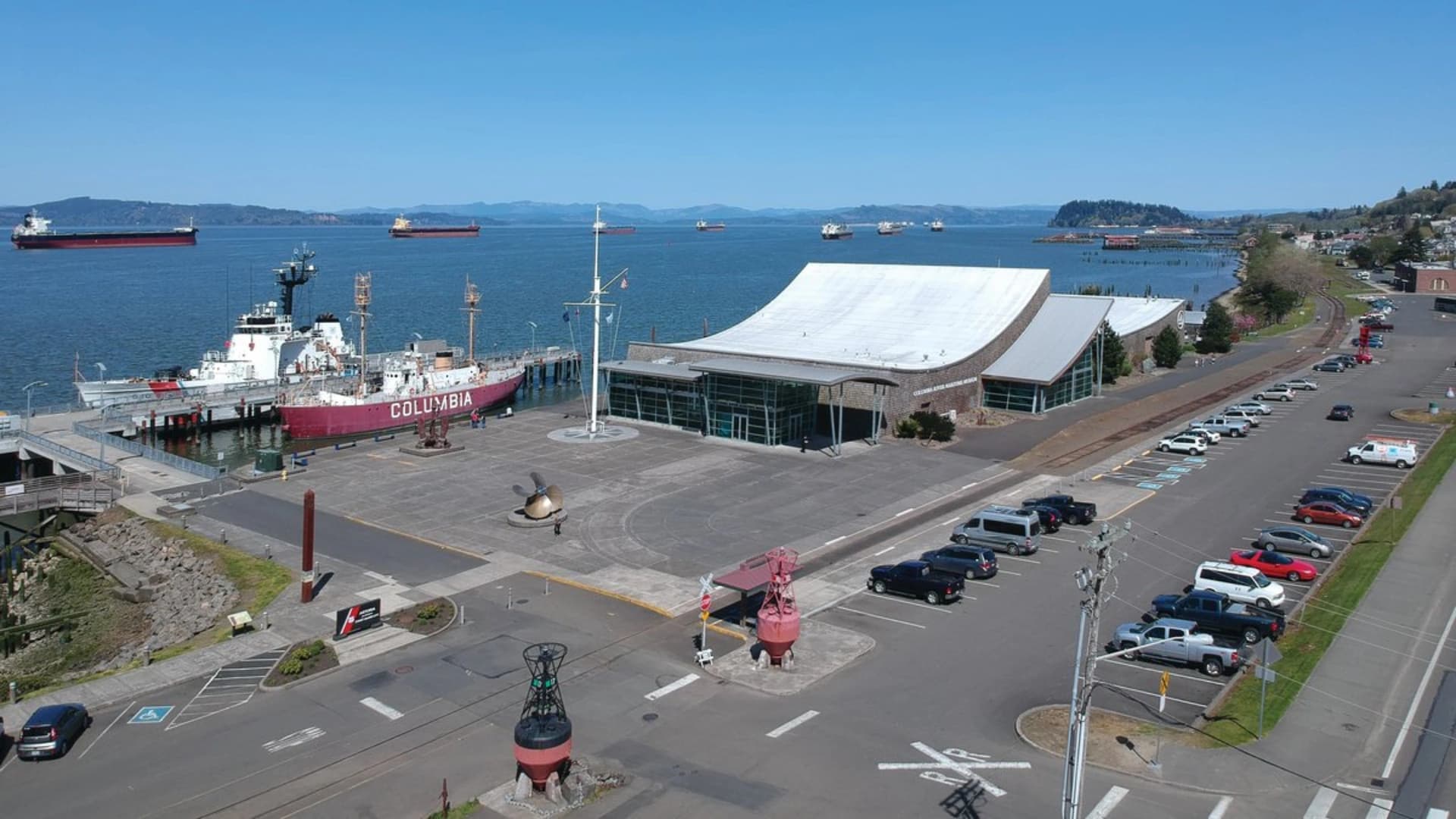 Aerial view of a waterfront area featuring a red ship named "Columbia" at dock, a modern building, and parked cars.