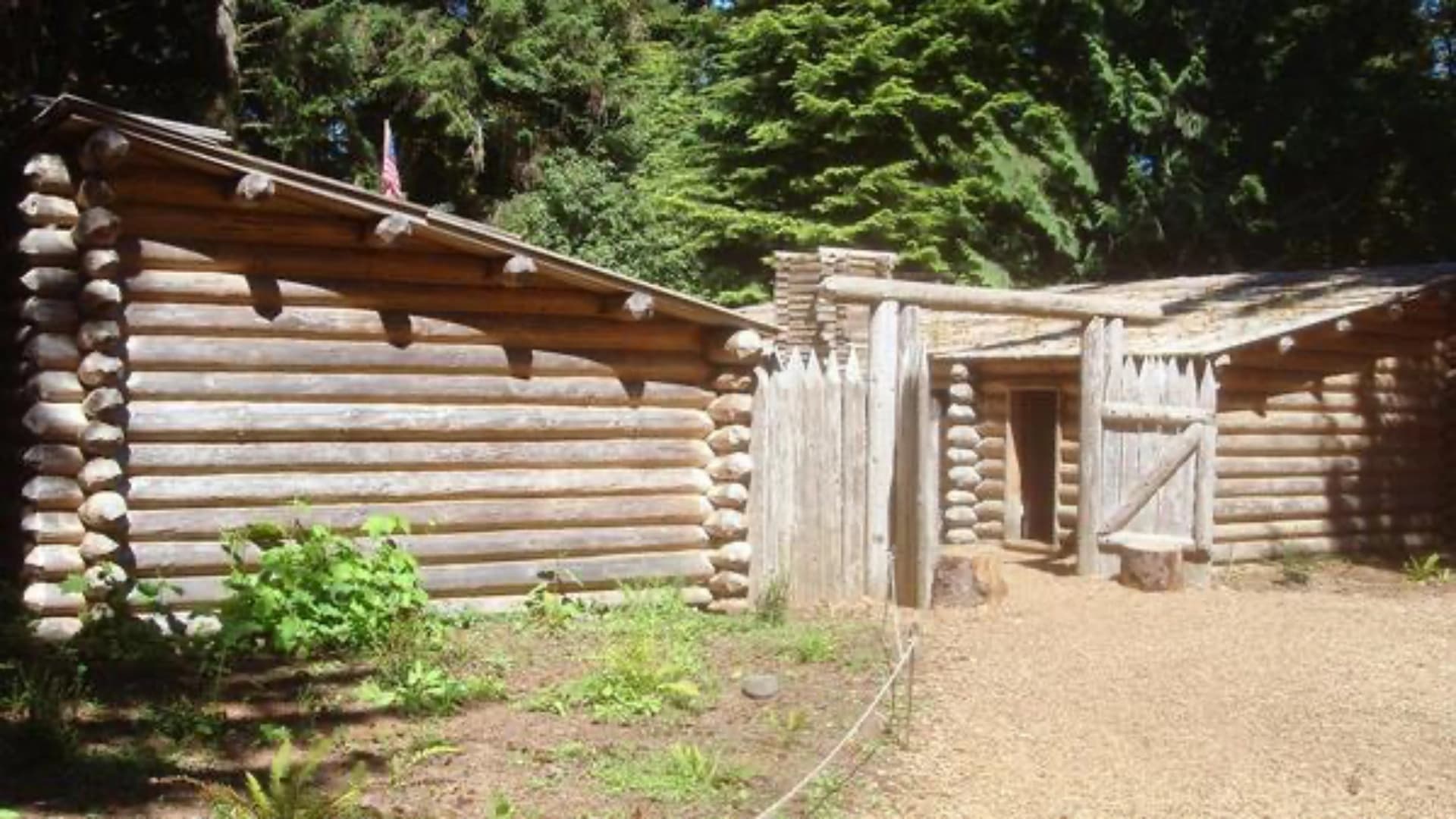 Log cabin structures surrounded by greenery, featuring a wooden fence and a gravel pathway.