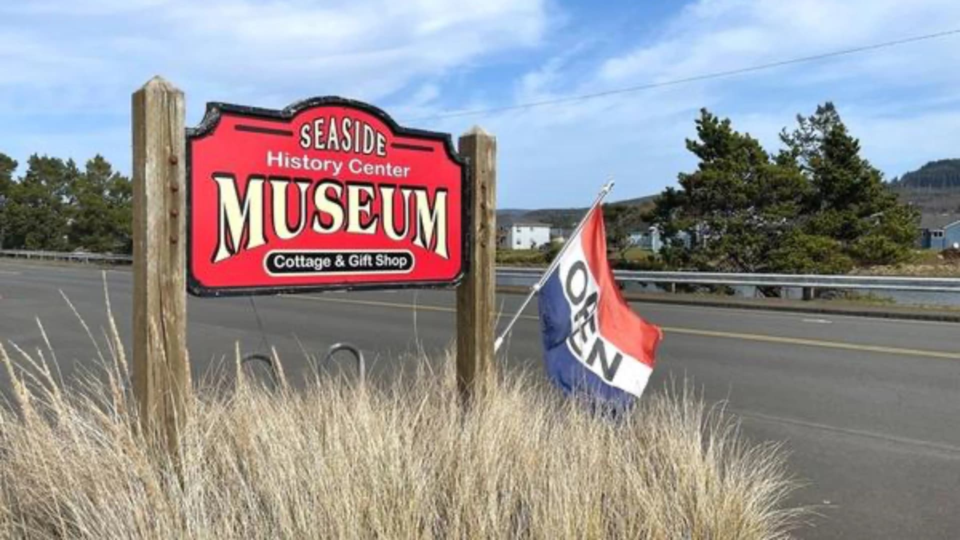 Sign for Seaside History Center Museum with "Open" flag, surrounded by tall grass and trees.