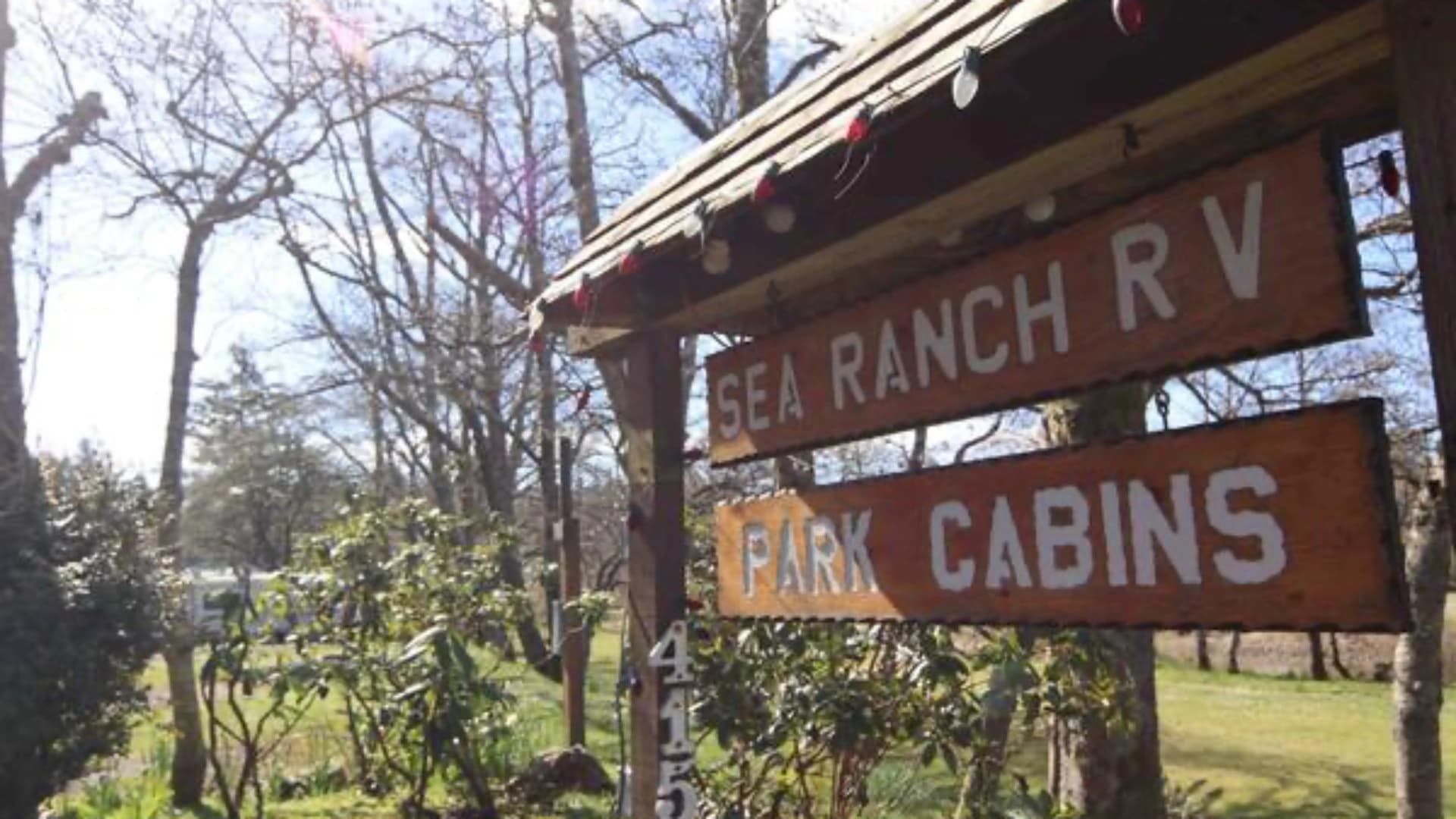A wooden sign for "Sea Ranch RV Park Cabins" surrounded by trees and greenery on a sunny day.