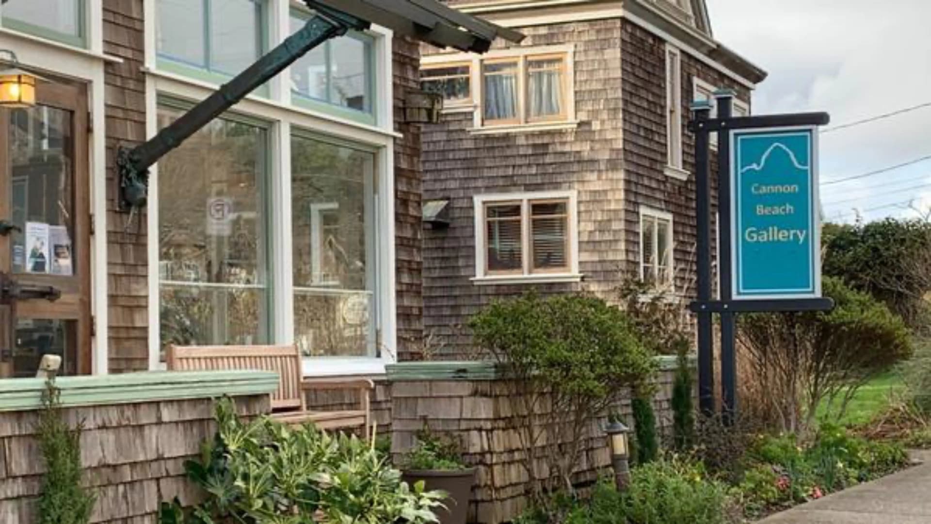 Exterior of the Cannon Beach Gallery, featuring wood paneling, a bench, and a sign in teal and white.