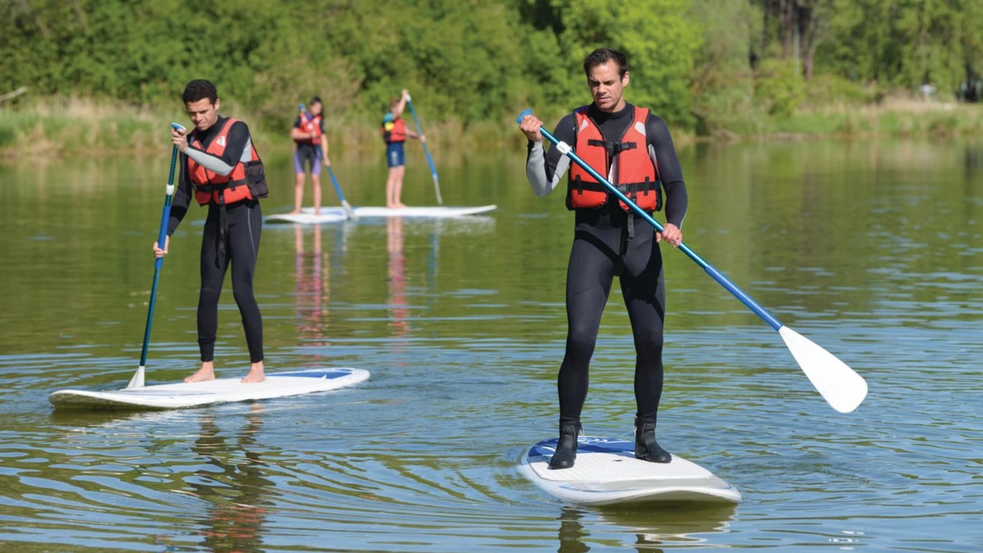Four people paddleboarding on a calm lake, wearing life jackets and wetsuits, surrounded by greenery.