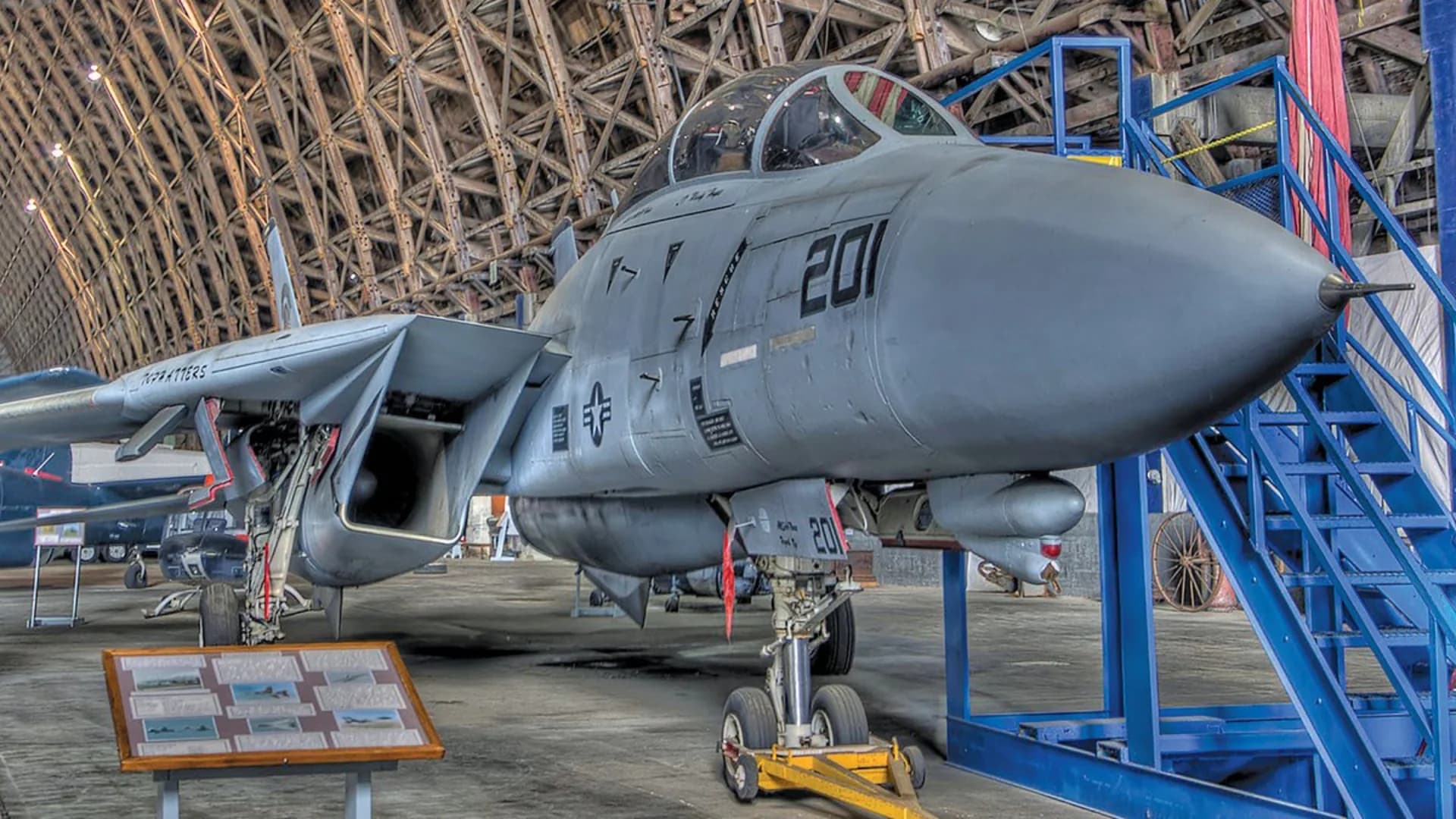Fighter jet displayed in a hangar, showcasing its fuselage and landing gear. Information plaque visible in front.