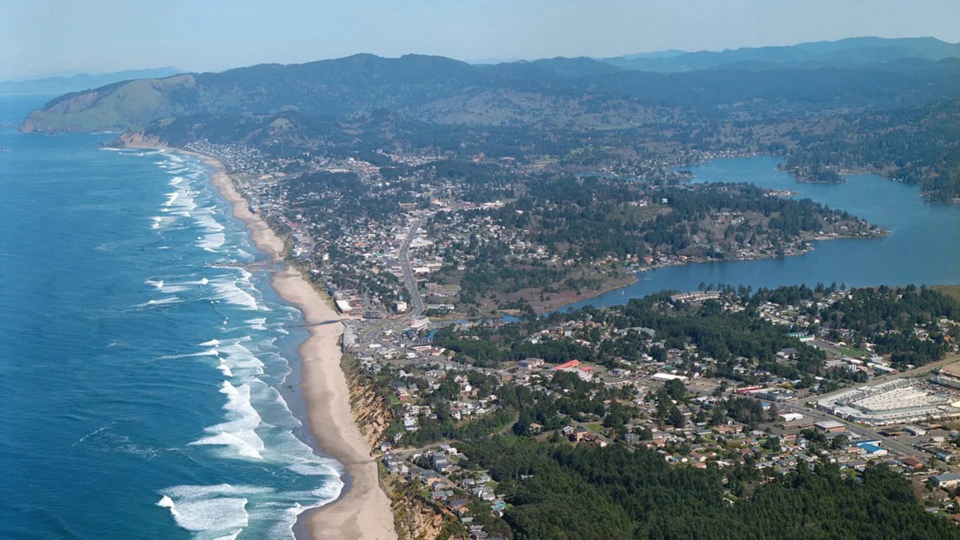 Aerial view of a coastal town with sandy beaches, waves, and surrounding hills and forested areas.