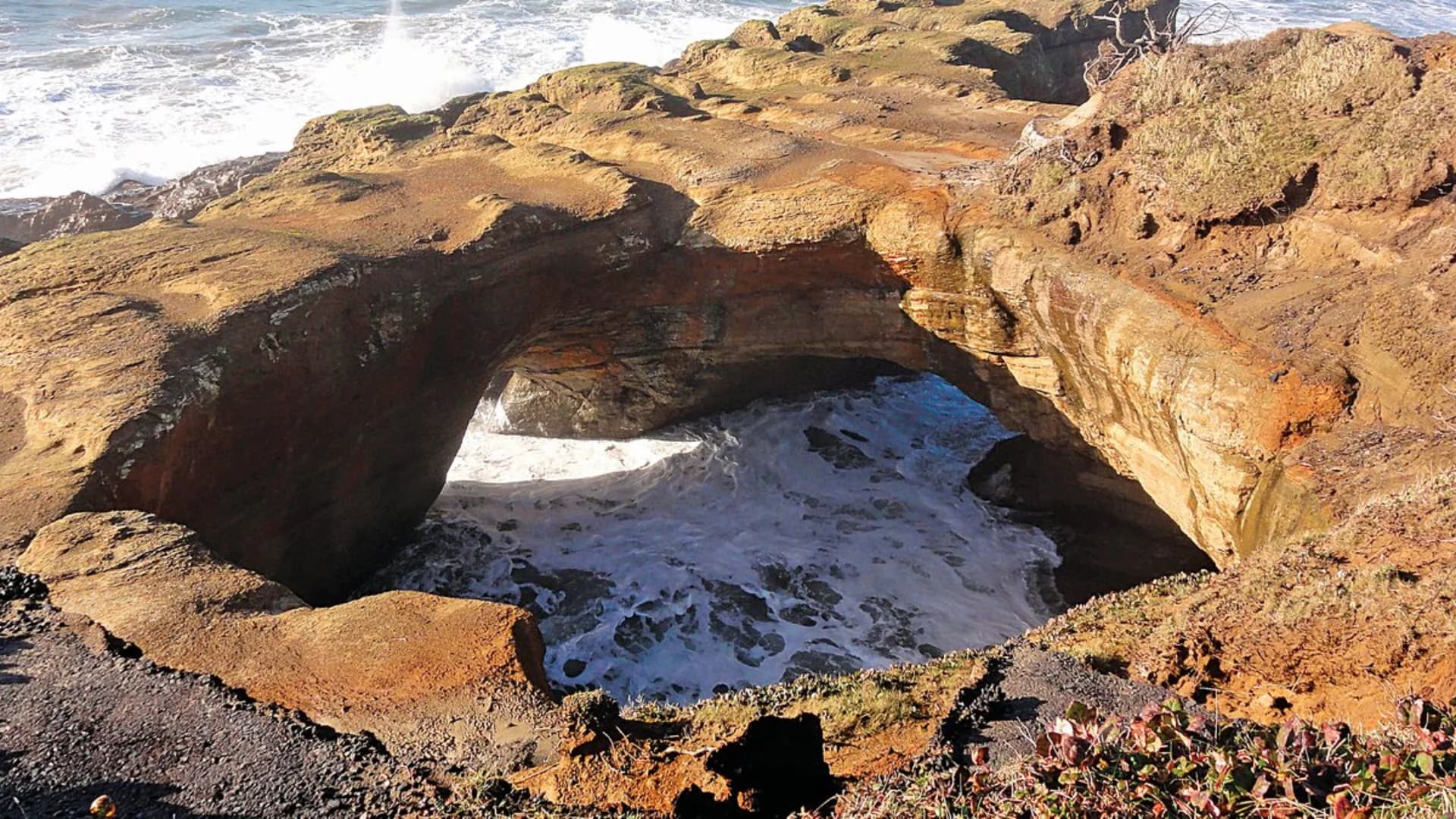 A natural rock arch above foamy ocean waves, framed by rugged coastal cliffs.