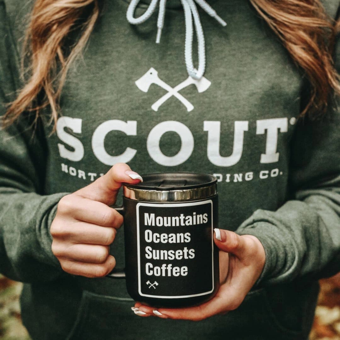 A person in a green sweatshirt holds a black mug with text: "Mountains Oceans Sunsets Coffee."