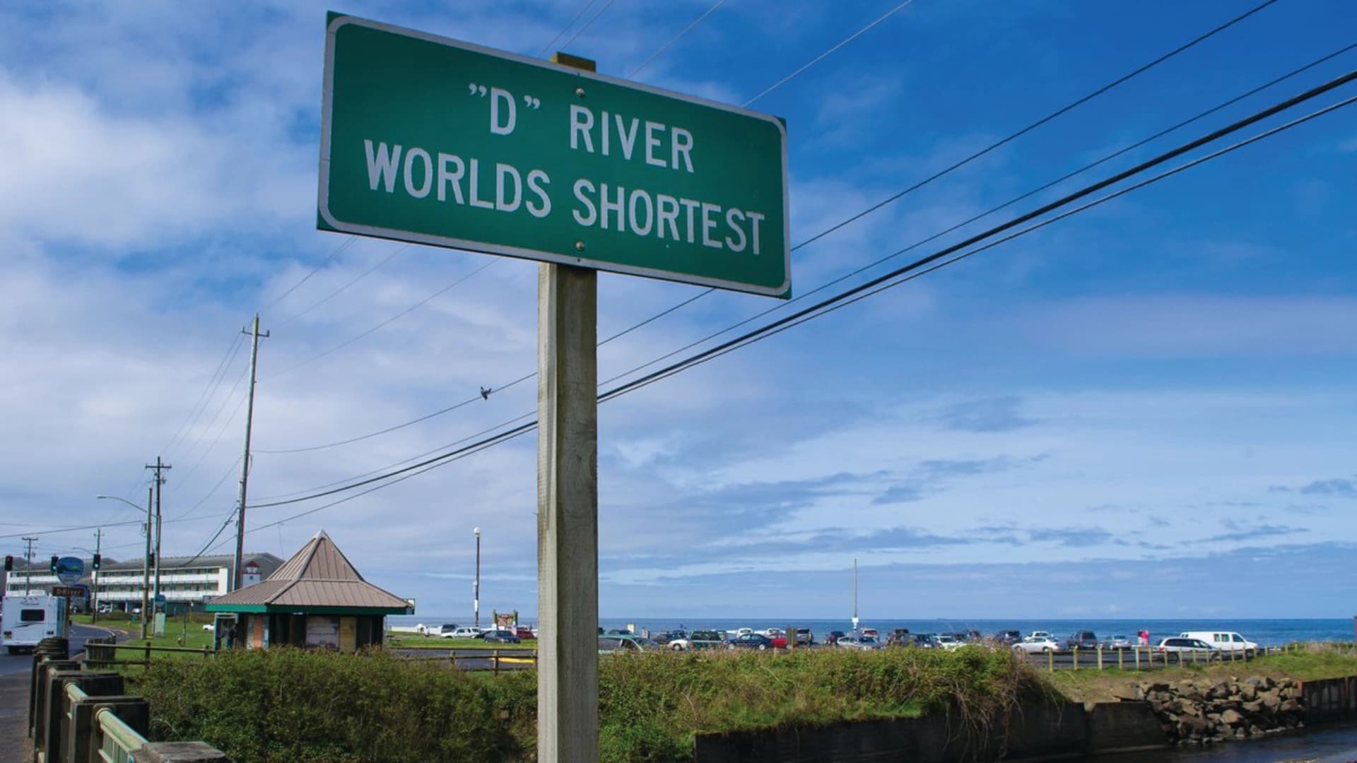 Signpost reading "D RIVER WORLDS SHORTEST" with a beach and ocean in the background under a blue sky.