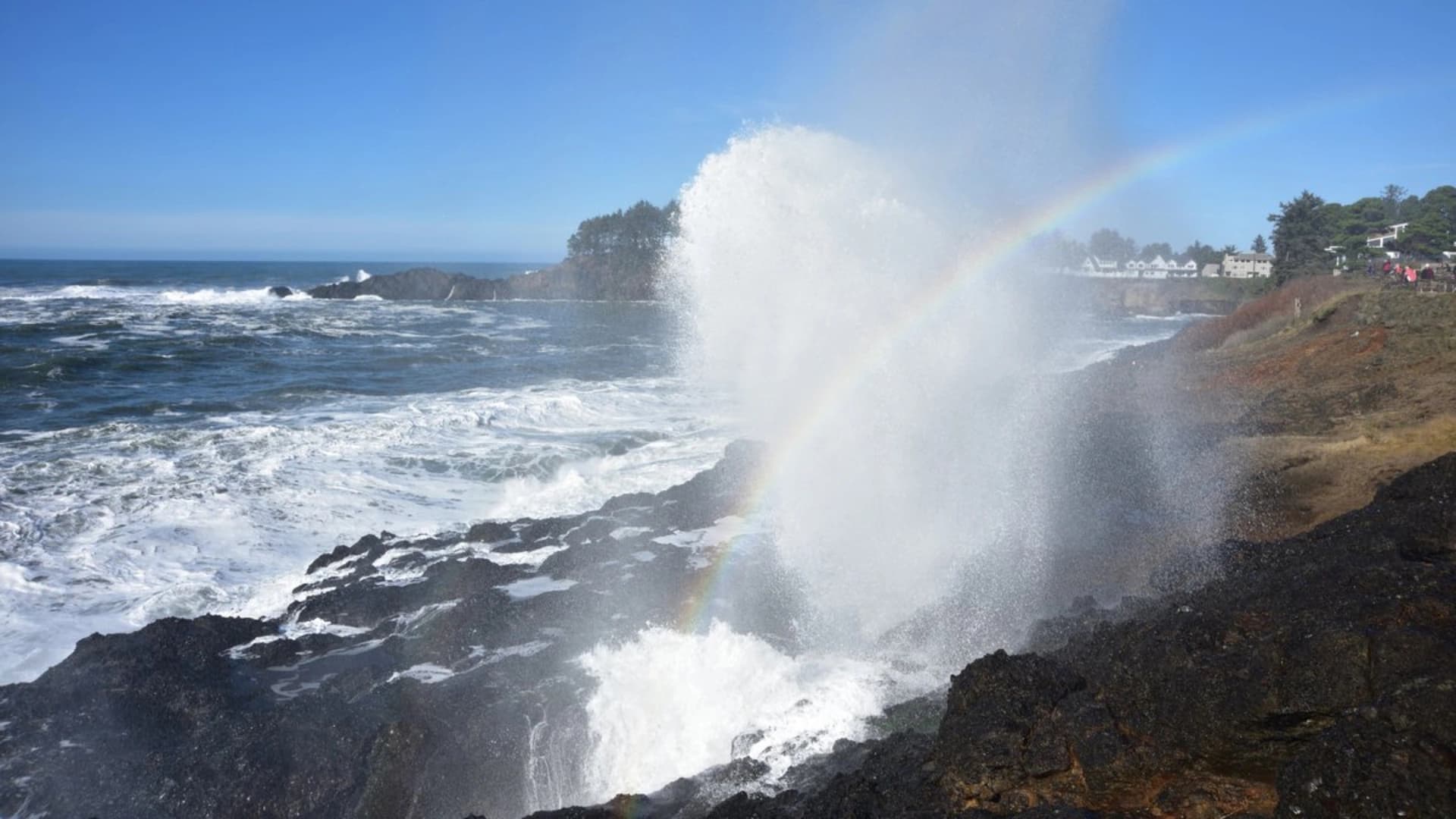 Waves crash against rocky shore with a spray creating a rainbow in the mist under a clear blue sky.
