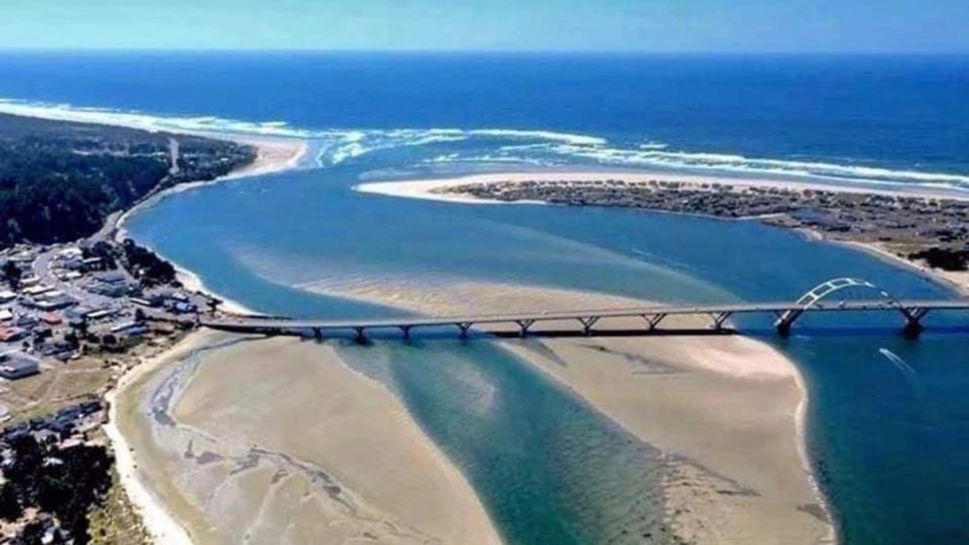 Aerial view of a bridge over a river, leading to coastal land with sandy beaches and ocean waves in the background.