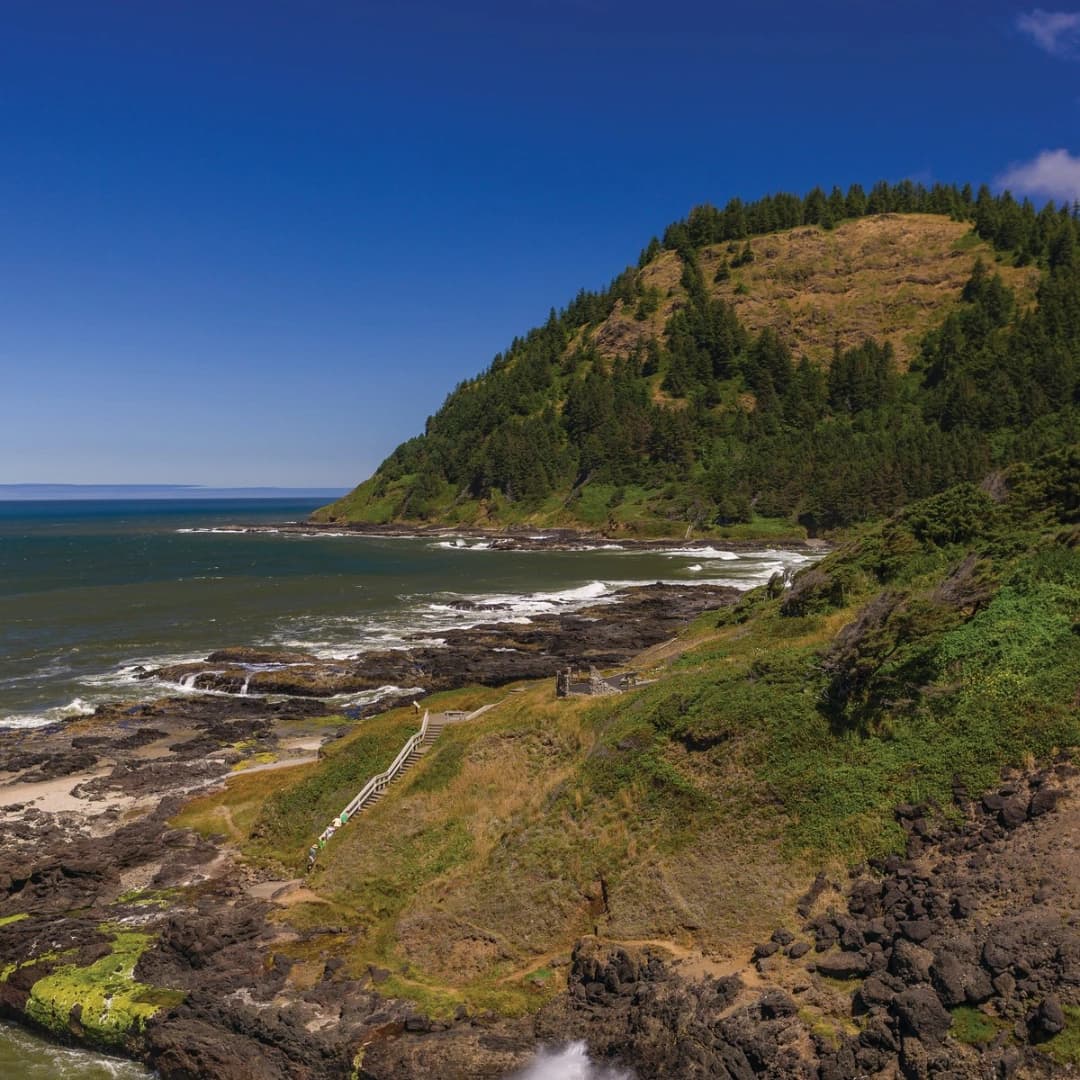 Coastal landscape with rocky shore, green hills, and a clear blue sky. Waves wash against the rocks.