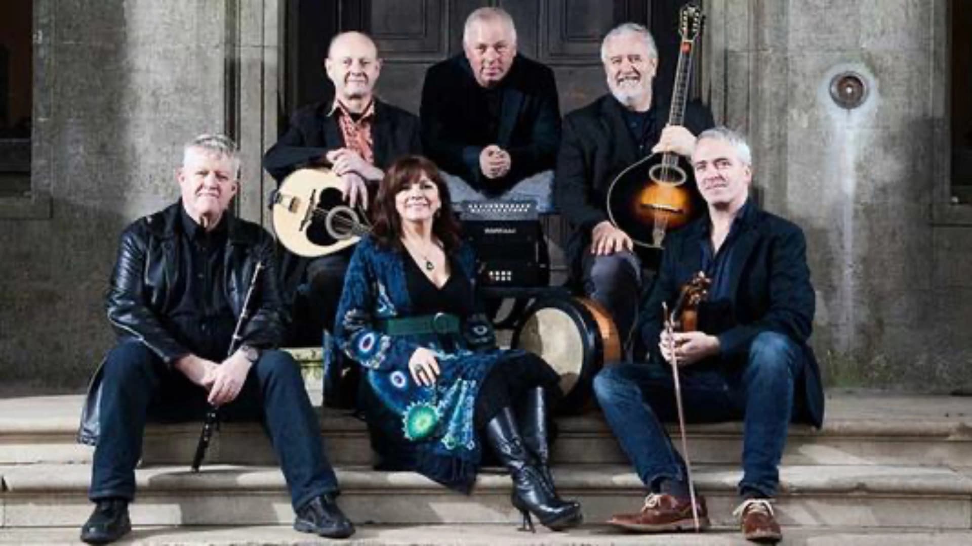 A band of six musicians posing on steps, holding various instruments like guitars and a flute.