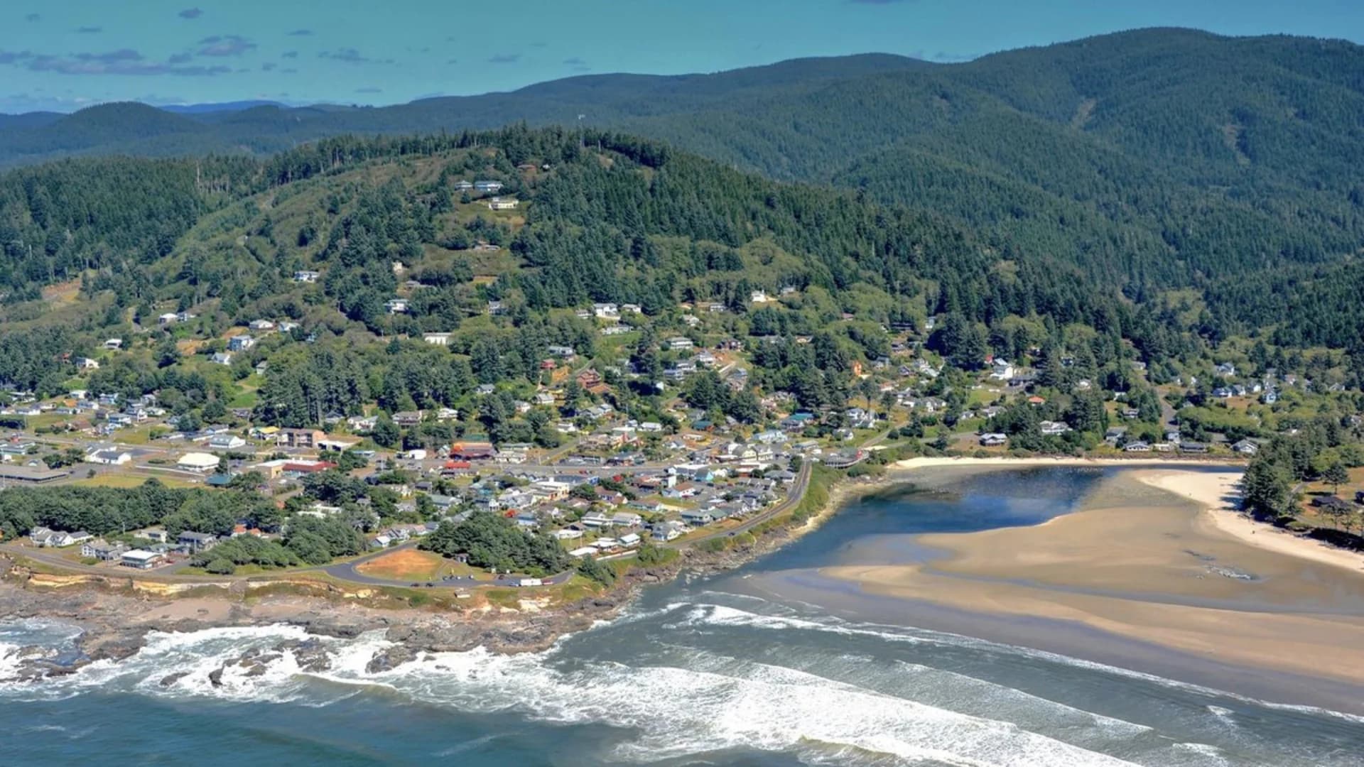 Aerial view of a coastal town nestled between green hills and sandy shores, with calm ocean waves nearby.