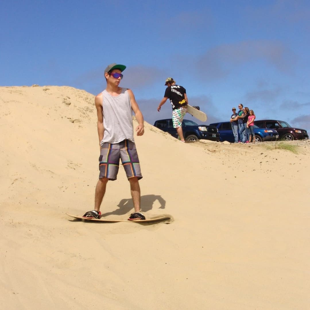 A young man stands on a sandboard on a dune, while another person walks behind him carrying a board.
