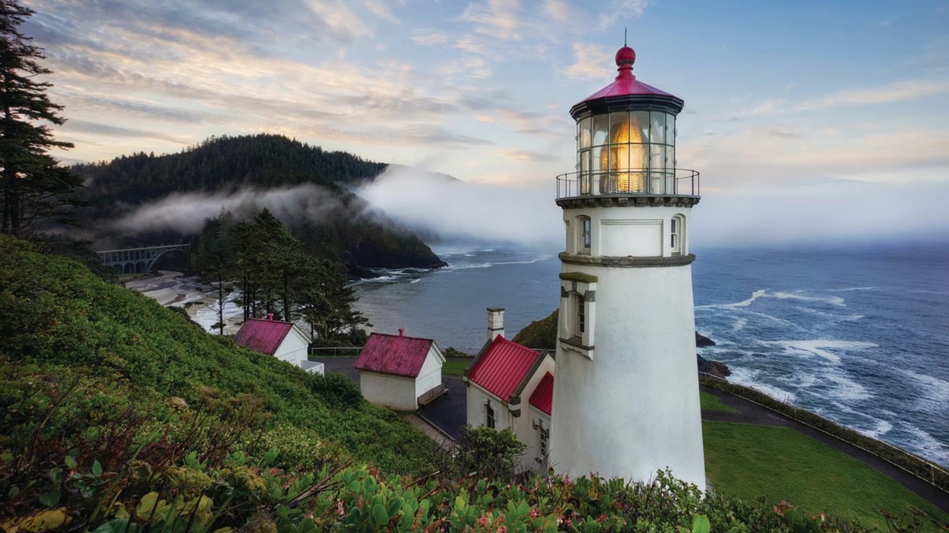 A lighthouse with a red roof sits on a cliff, overlooking a misty coastline with waves and a bridge in the distance.