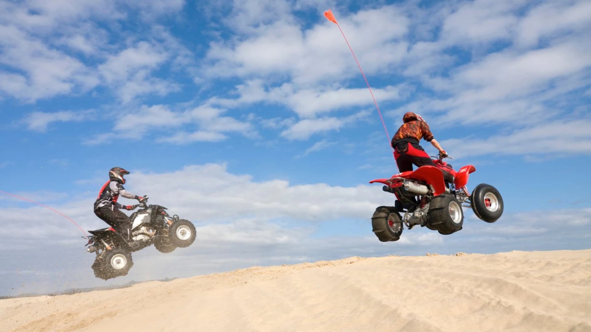 Two riders perform jumps on all-terrain vehicles (ATVs) over sandy terrain under a blue sky.