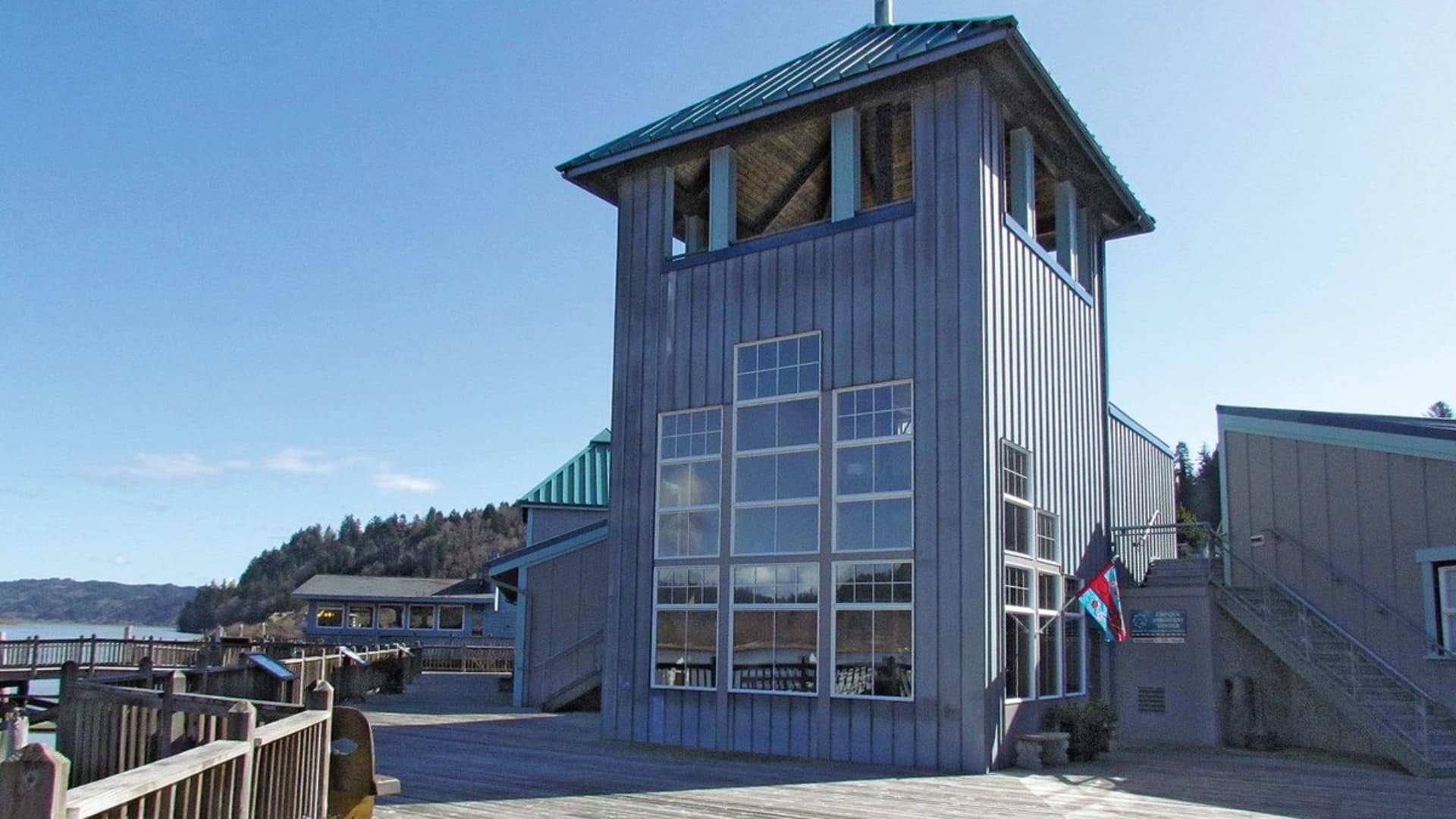 Gray wooden building with large windows and a teal roof, situated near a waterfront boardwalk.