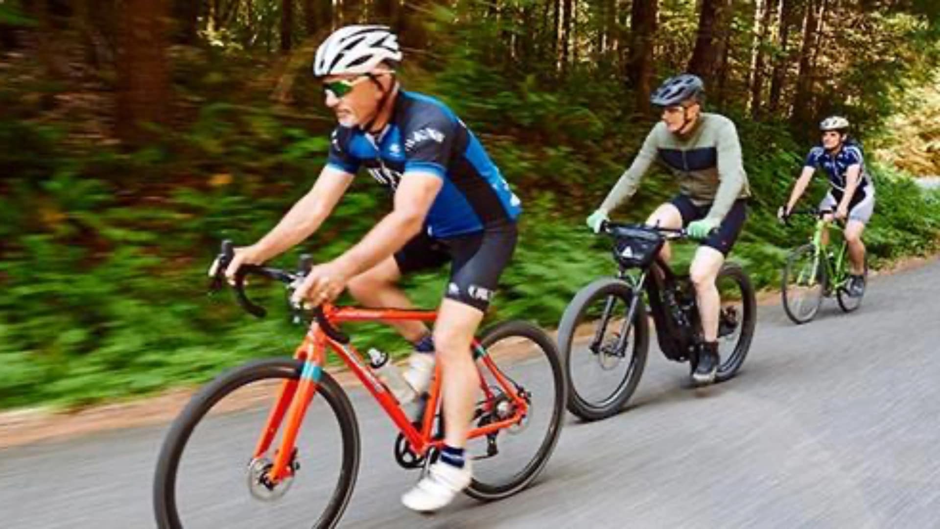 Three cyclists ride on a paved path through a lush, green forest.