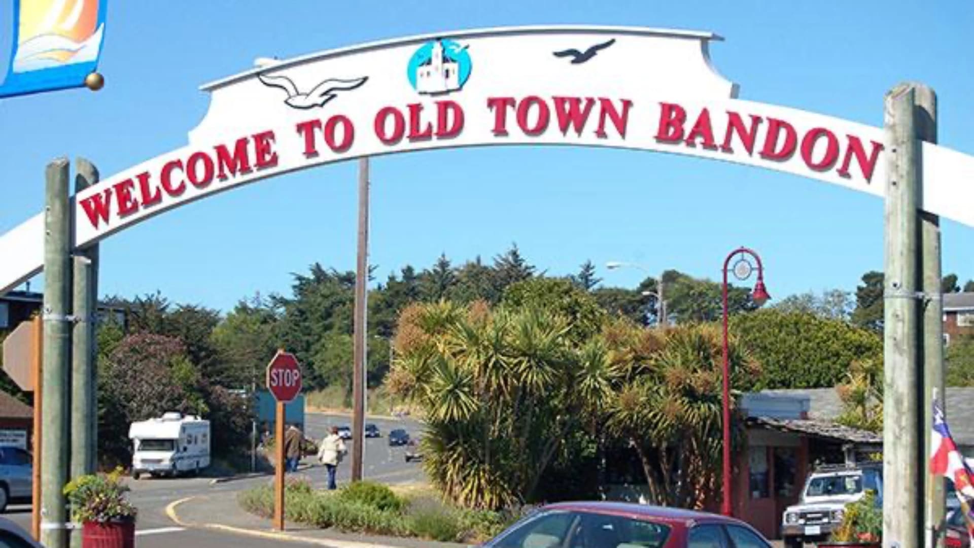 Archway sign reading "Welcome to Old Town Bandon" with palm trees and traffic in the background.
