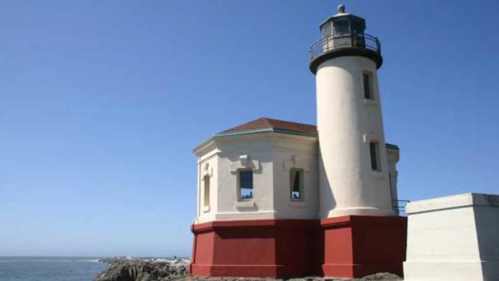 A white and red lighthouse stands by the shore under a clear blue sky.