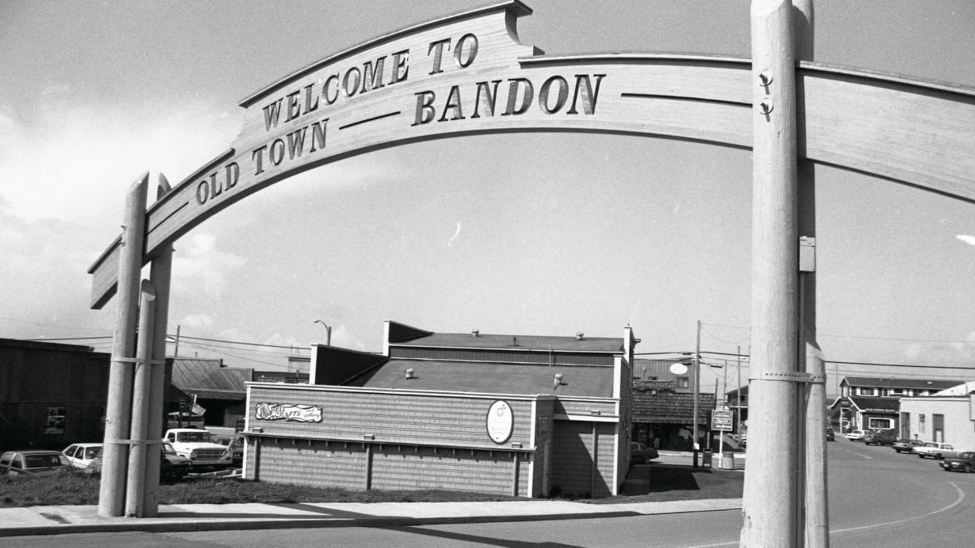 Wooden sign reading "Welcome to Old Town Bandon" above a street scene with buildings and parked cars.