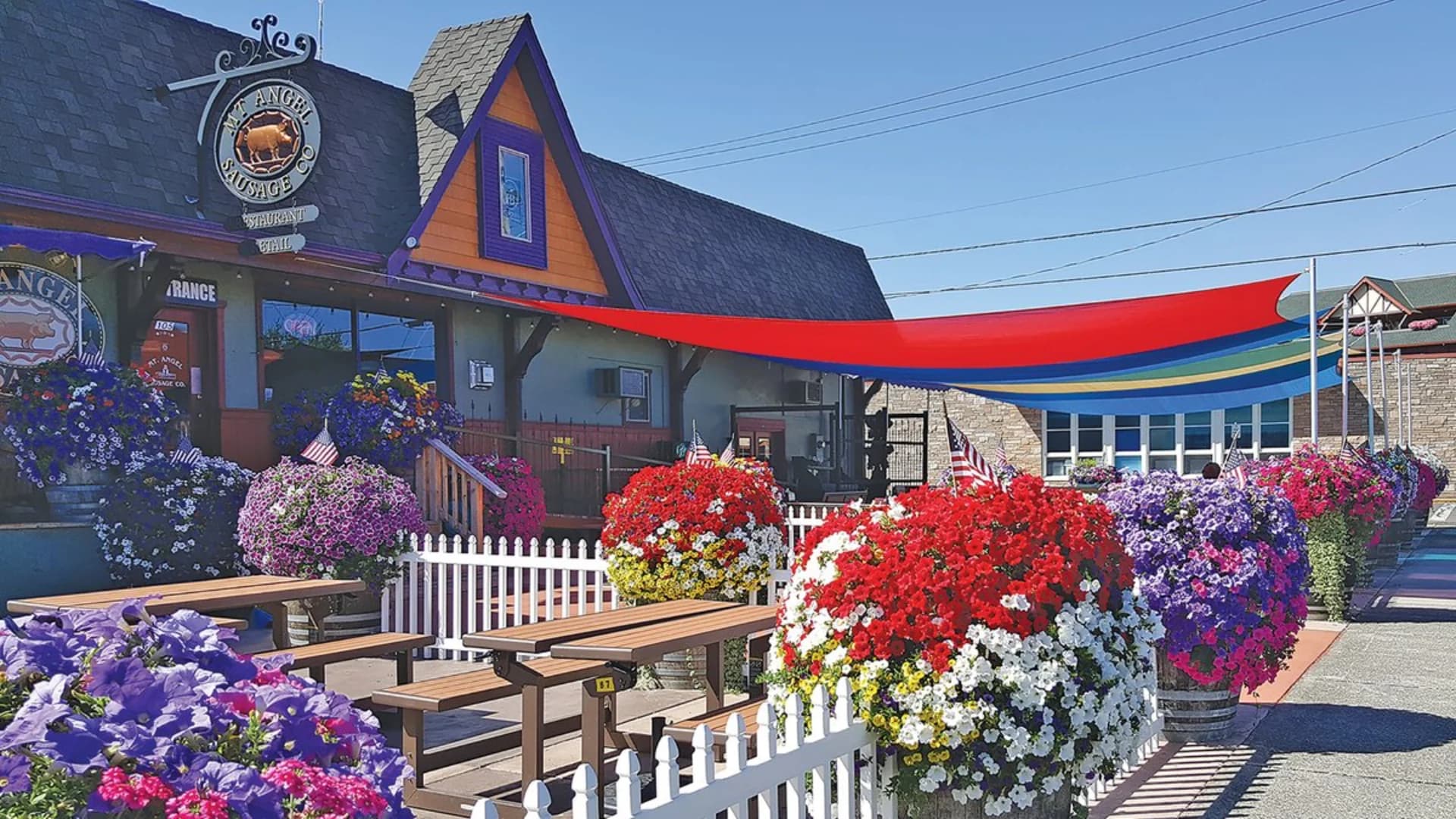 Colorful flowers in planters line the outdoor seating area of Mt. Angel Sausage Co. restaurant with vibrant awnings above.