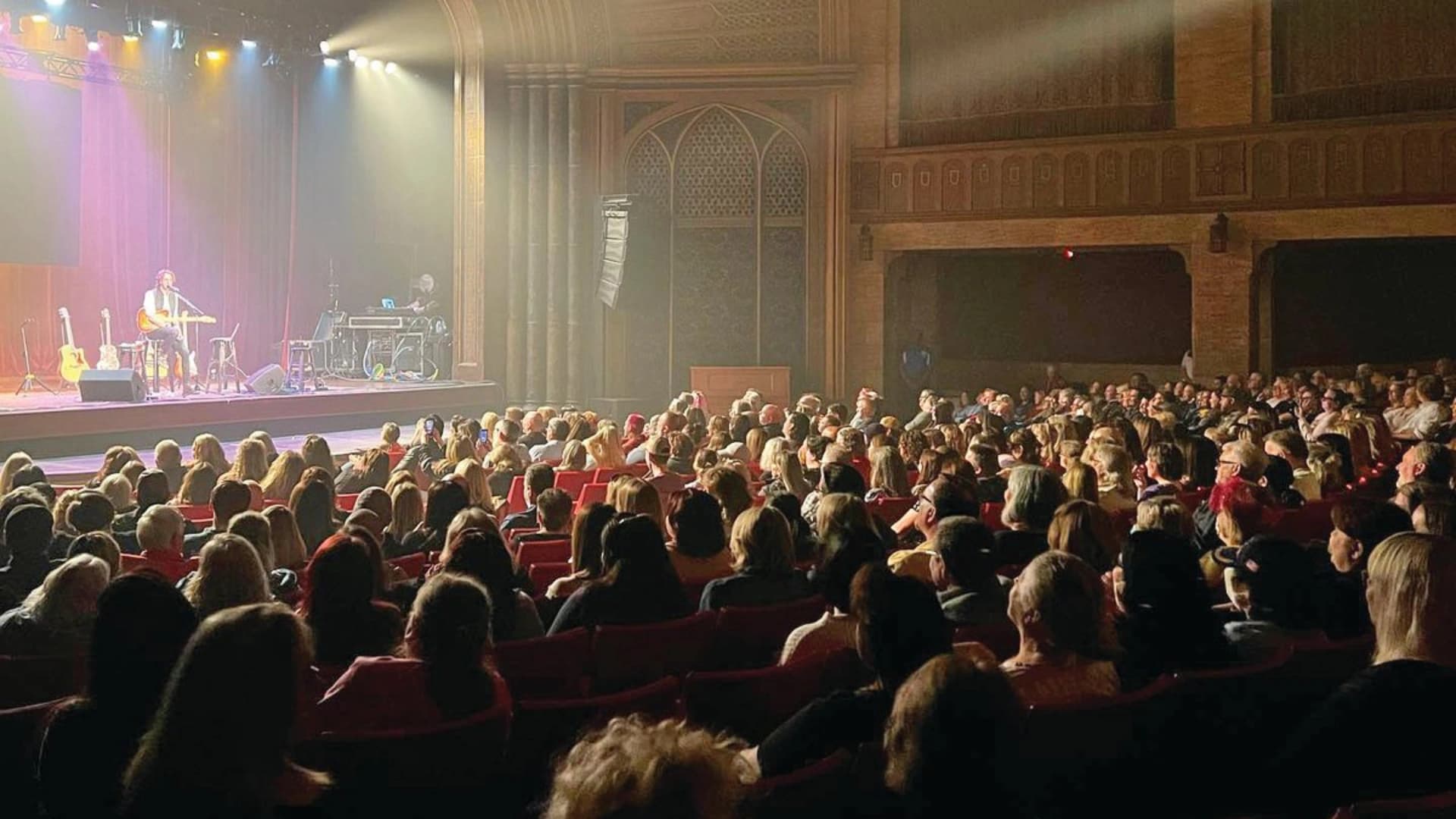 A musician performs on stage in a theater, with an audience seated in red chairs, eagerly watching the show.