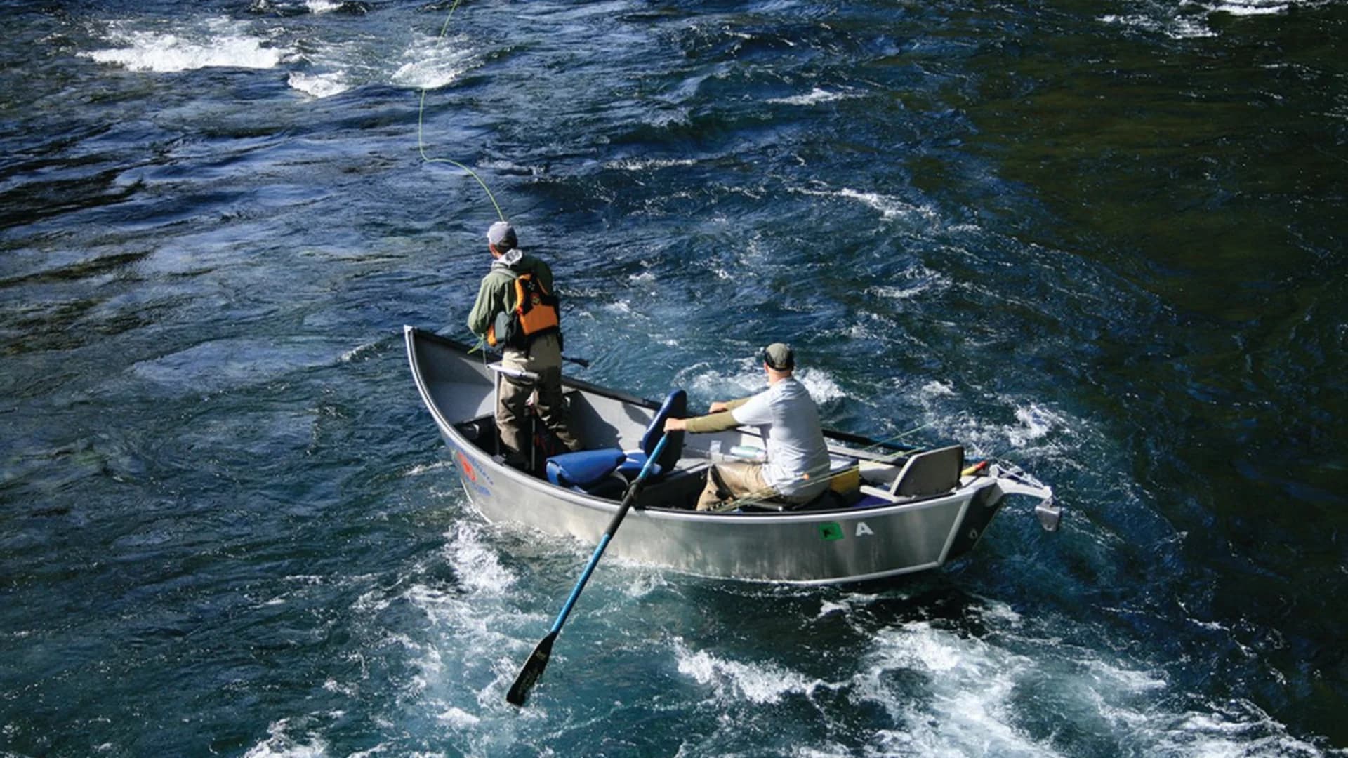 Two fishermen in a boat casting lines into a flowing river. Water reflects sunlight.