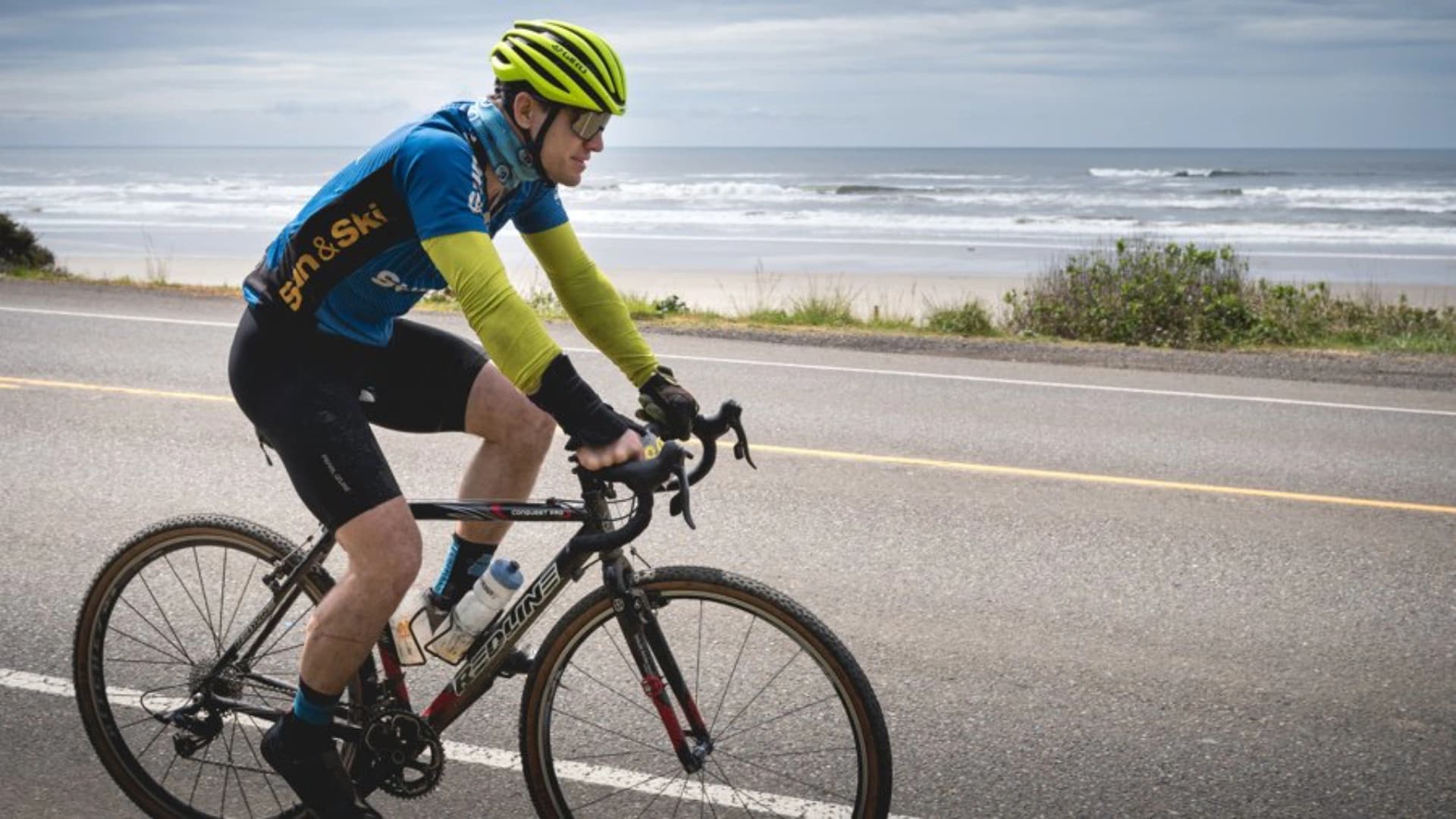 A cyclist in a blue and yellow jersey rides along a coastal road near the ocean.