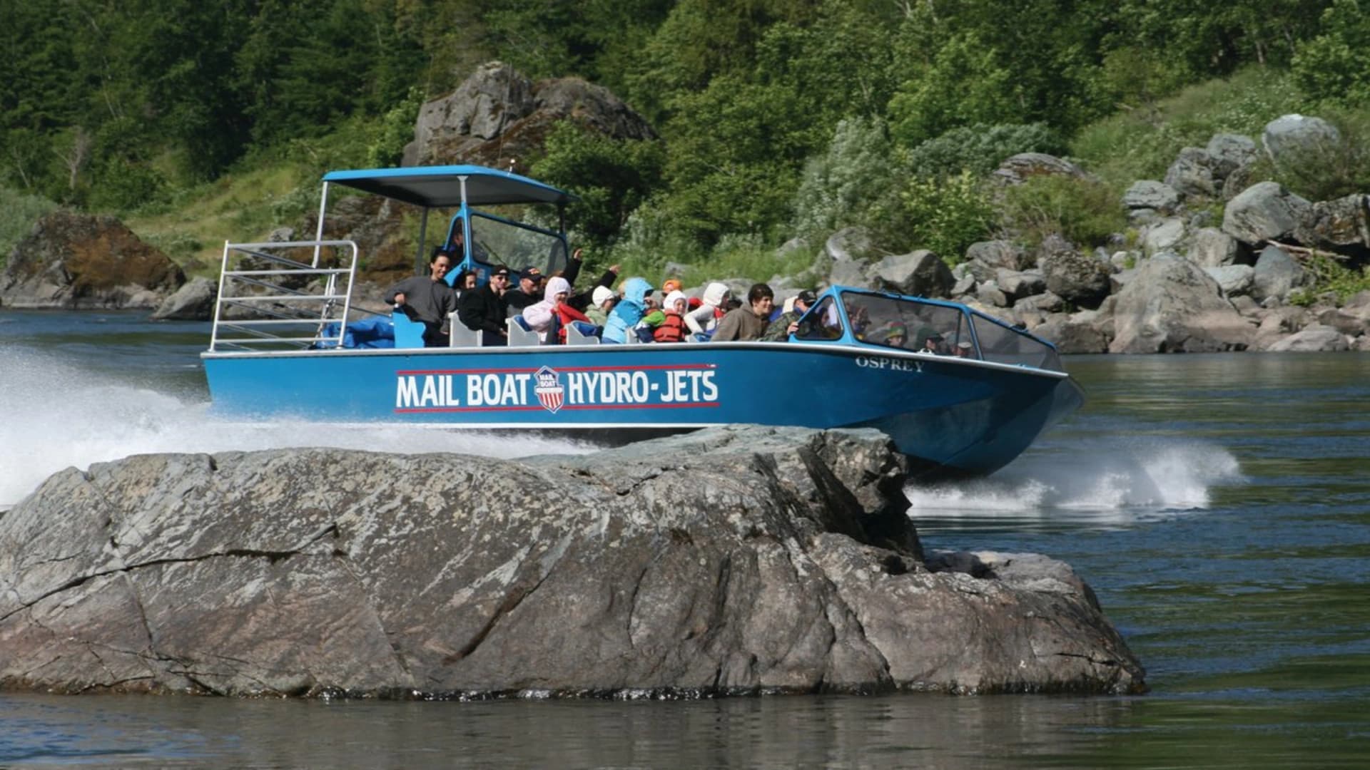A blue hydro-jet boat with passengers speeds past a large rock in a river, surrounded by greenery.