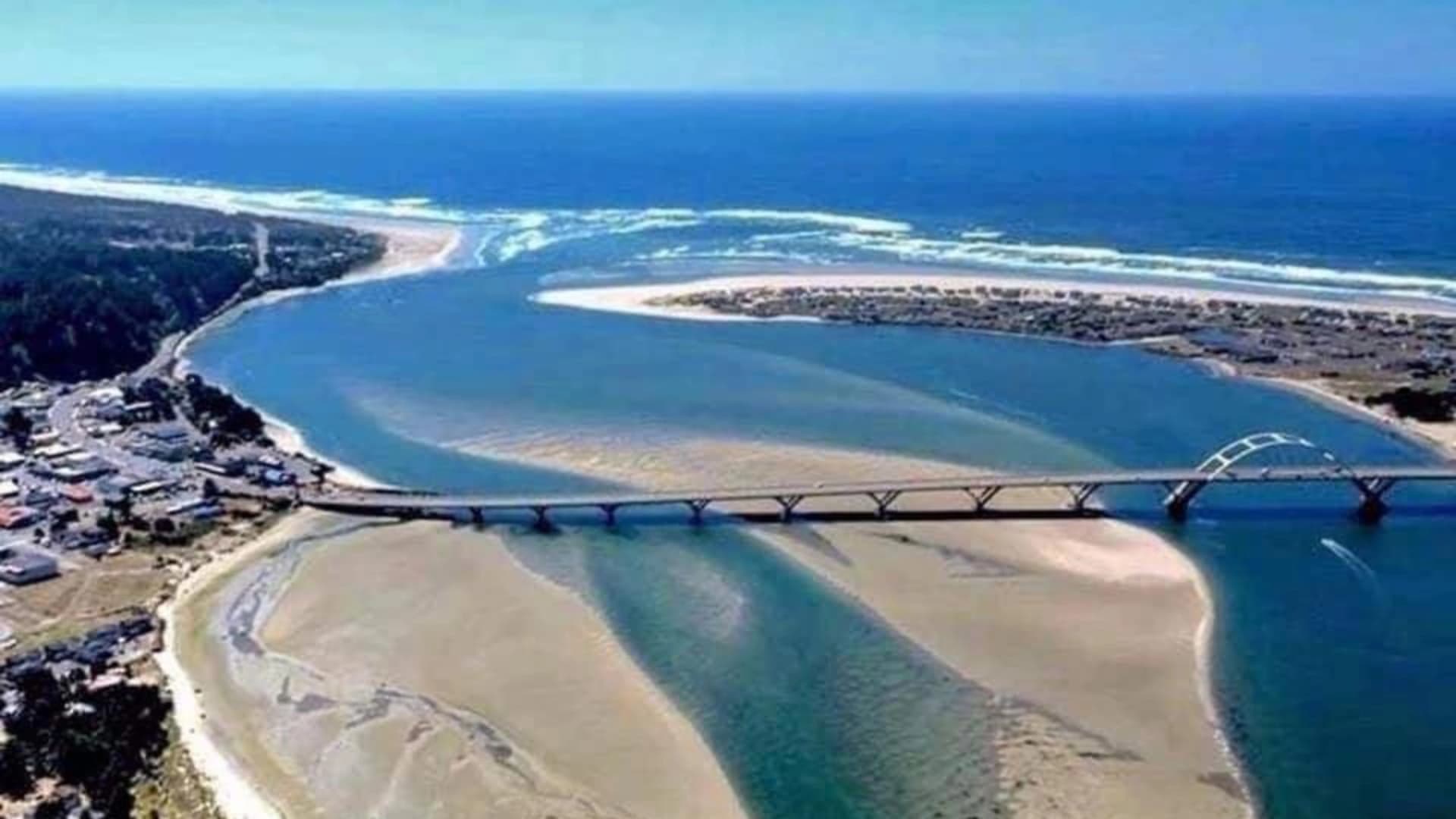 Aerial view of a river leading to the ocean, featuring a bridge and sandy shores.