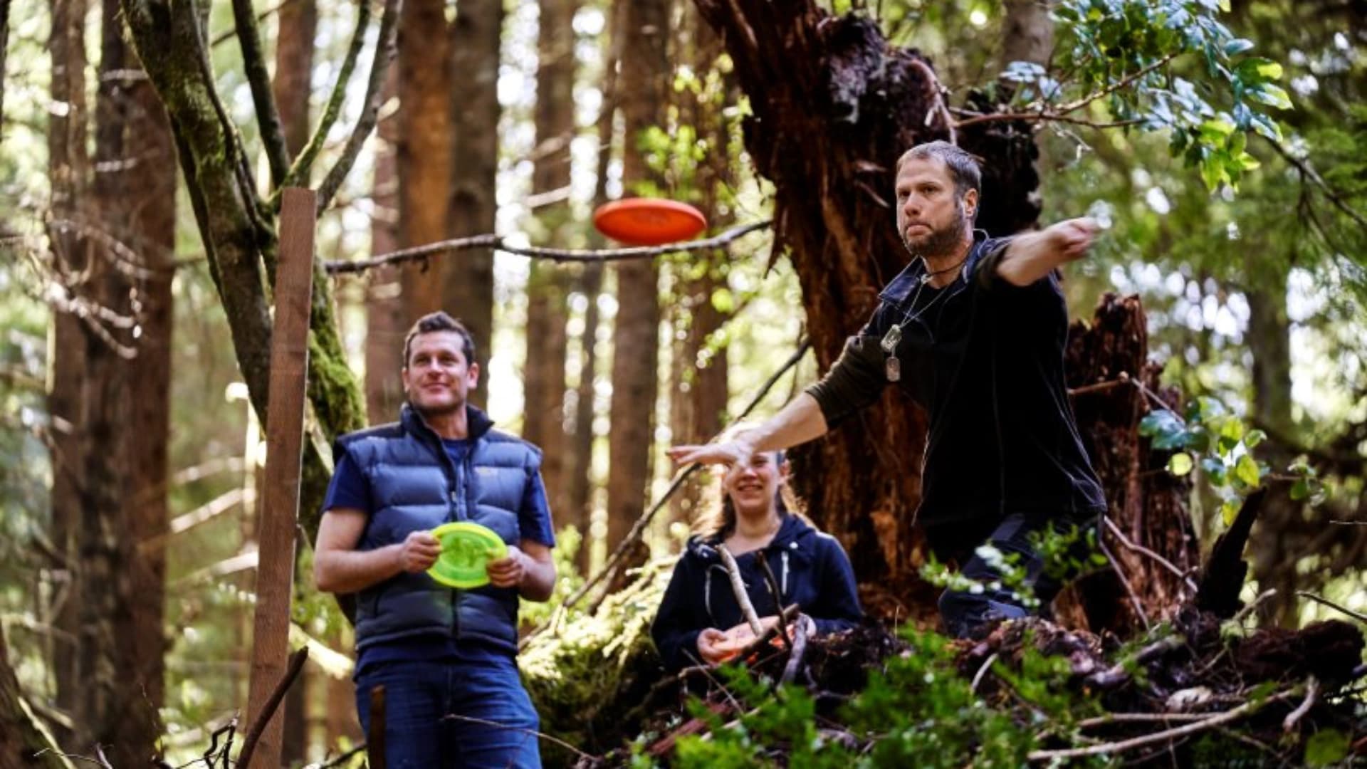 Three people enjoying disc golf in a forest; one is throwing a disc while the others watch.