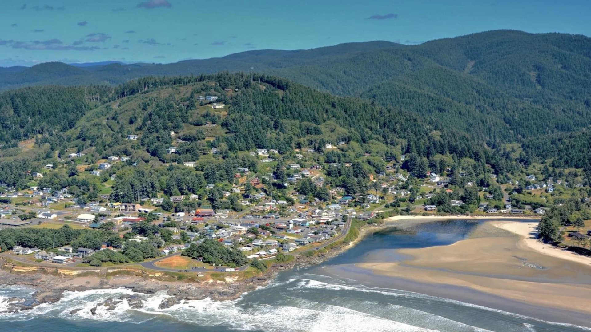 Aerial view of a coastal town surrounded by hills and trees, with waves crashing on the shore.