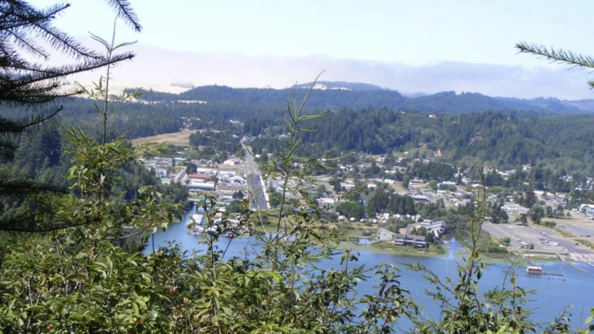A view of a small town beside a river, surrounded by trees and rolling hills under a clear sky.