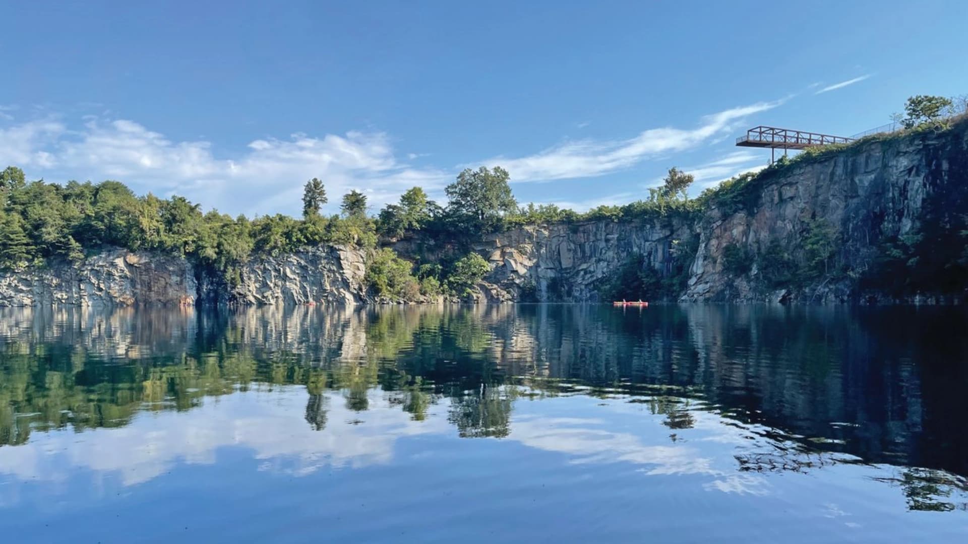 Serene quarry lake surrounded by trees and steep rocky cliffs, with a wooden overlook and two kayakers in the water.