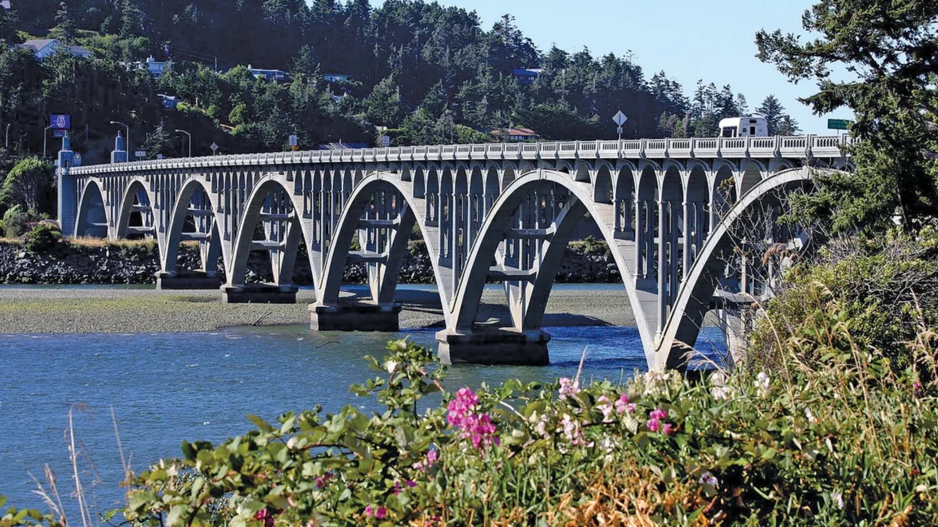 Concrete arch bridge spans a calm river, surrounded by trees and colorful flowers in the foreground.