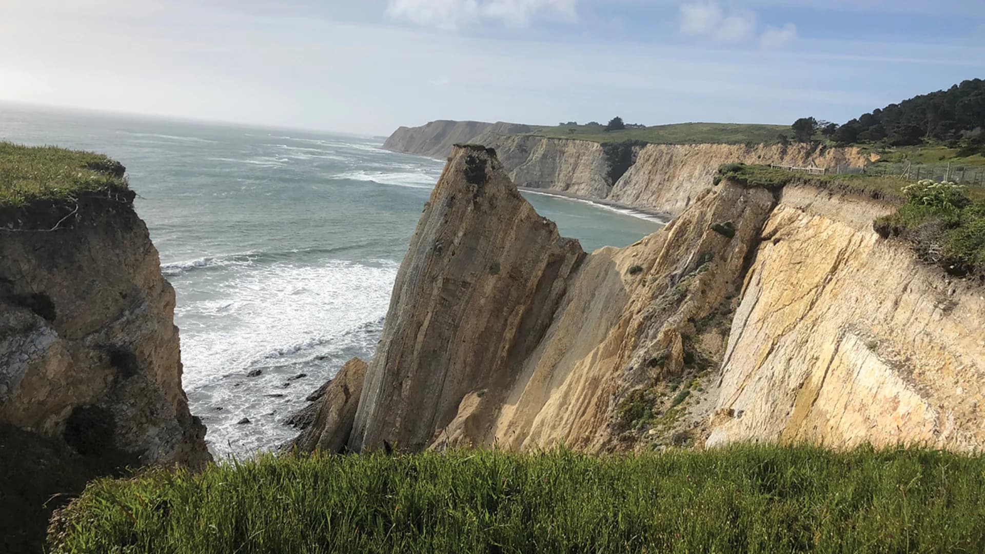 A dramatic coastal cliff with steep rock formations overlooking the ocean and distant shoreline under a blue sky.