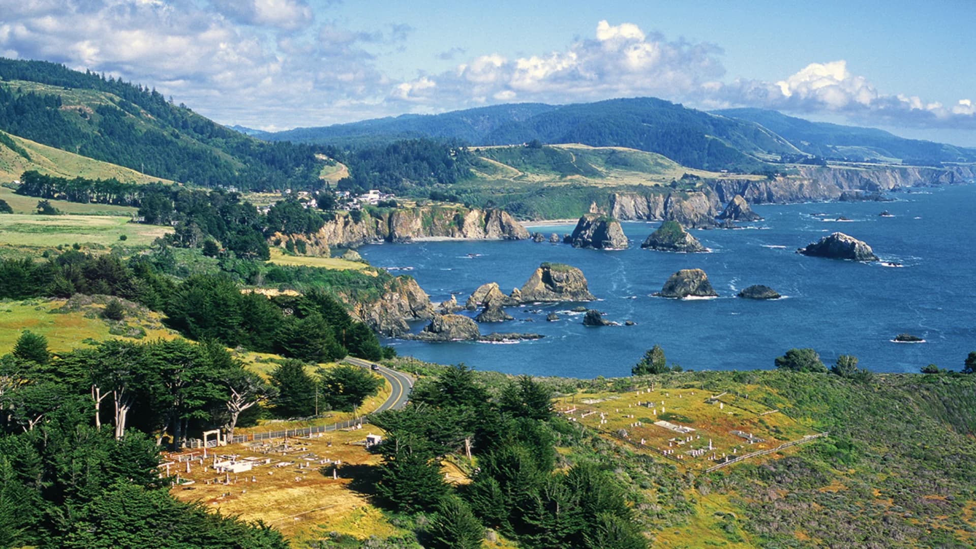 Coastal landscape with rocky cliffs, green hills, and a winding road along the shore under a partly cloudy sky.