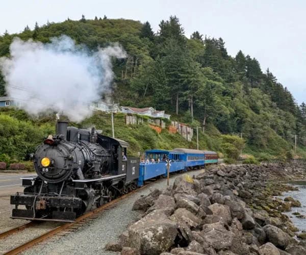 Steam locomotive pulls a blue passenger train along a track beside rocky shore and forested hillside.
